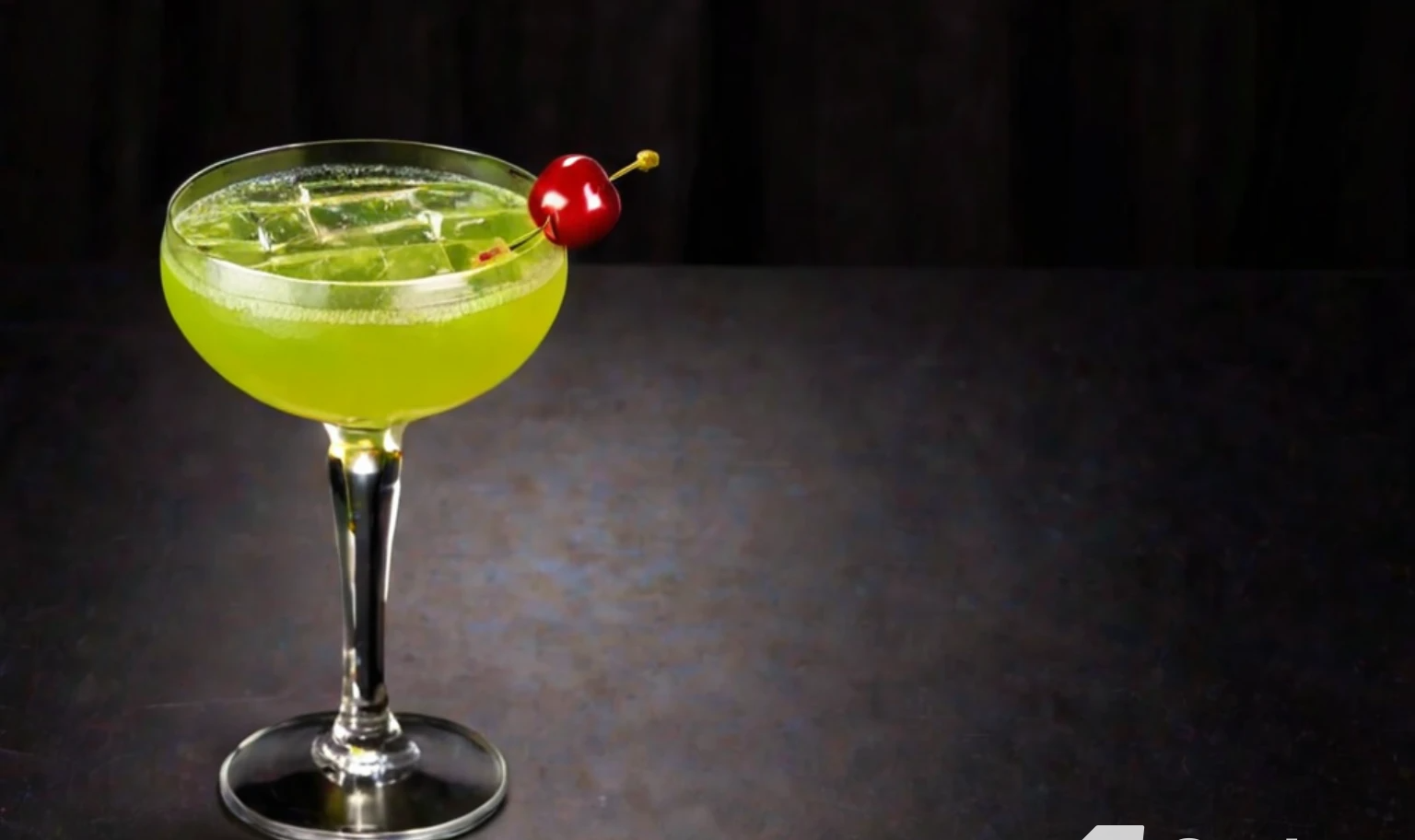 A glass of Last Word cocktail garnished with a lime twist, with bottles of gin, green chartreuse, maraschino liqueur, and fresh limes arranged around it.