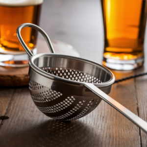“Close-up of a stainless steel bar strainer, featuring a handle and a perforated bowl designed to filter out ice and other solids from cocktails. The strainer is positioned against a clean, modern bar surface, showcasing its polished finish and practical design.”